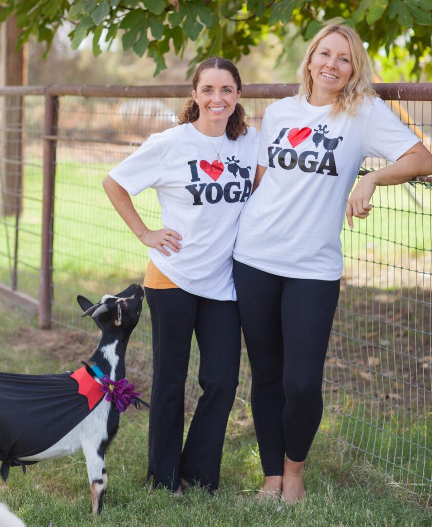Two girls April Gould and Sarah Williams who are the original creators and first entrepreneurs of goat yoga leaning against a fence with a goat looking up at them wearing an I love goat yoga shirts in the Gilbert Arizona sunset 2015
