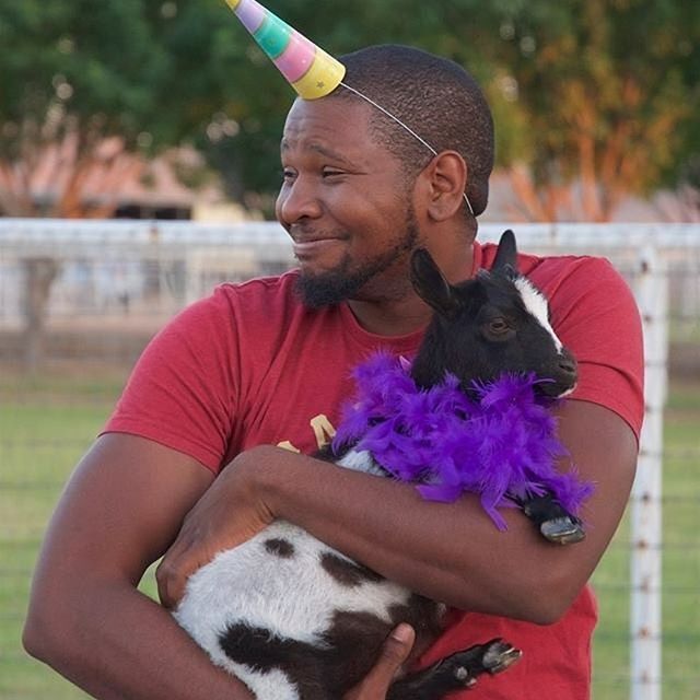 a happy African American man at the first goat yoga in Gilbert Arizona smiling while holding a black and white baby goat with a party rainbow unicorn horn on his head looking at the original creators, April Gould and Sarah Williams who started goat yoga in 2015.