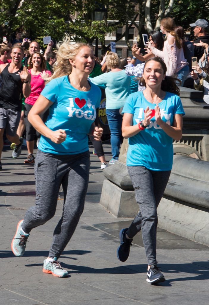 April Gould and Sarah Williams running to the starting line of The Amazing Race season 30  in blue goat yoga shirts while cheering through a crowd of people. 