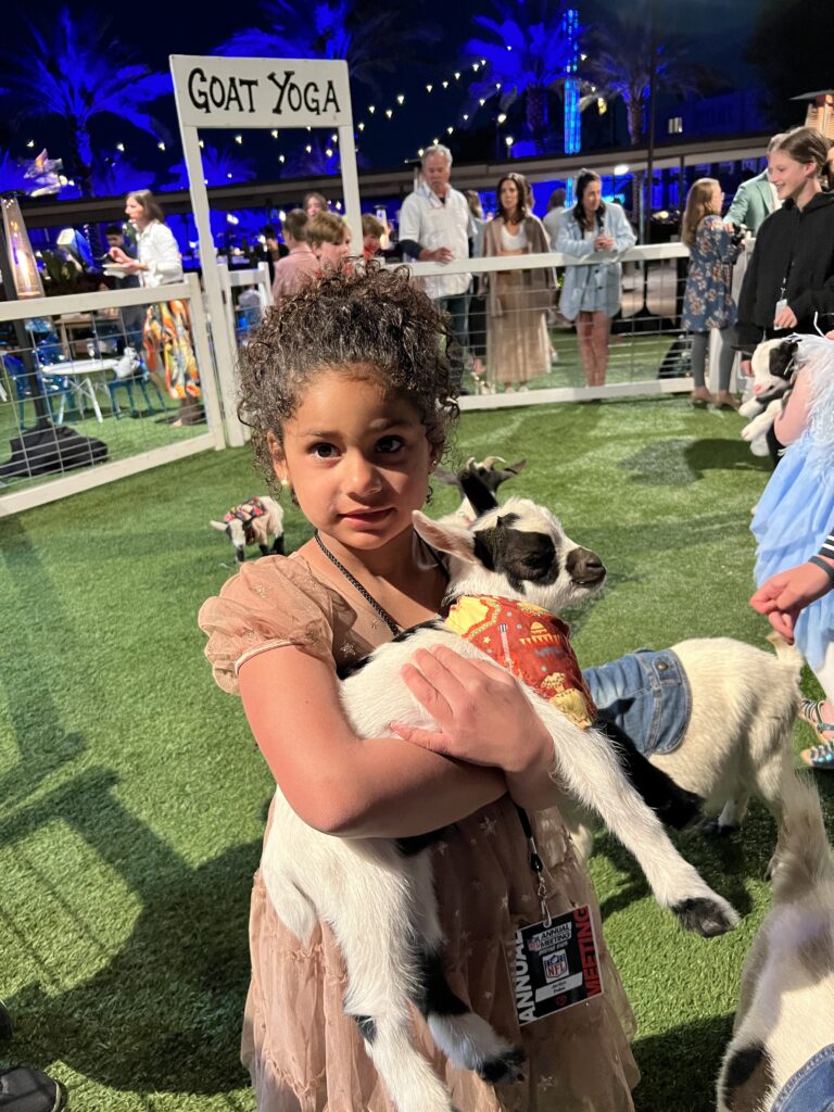 Beautiful African American girl with her hair in a curly ponytail and pink dress holding a baby goat under his belly in front of a sign that says goat yoga at a party in the Arizona Sunset.